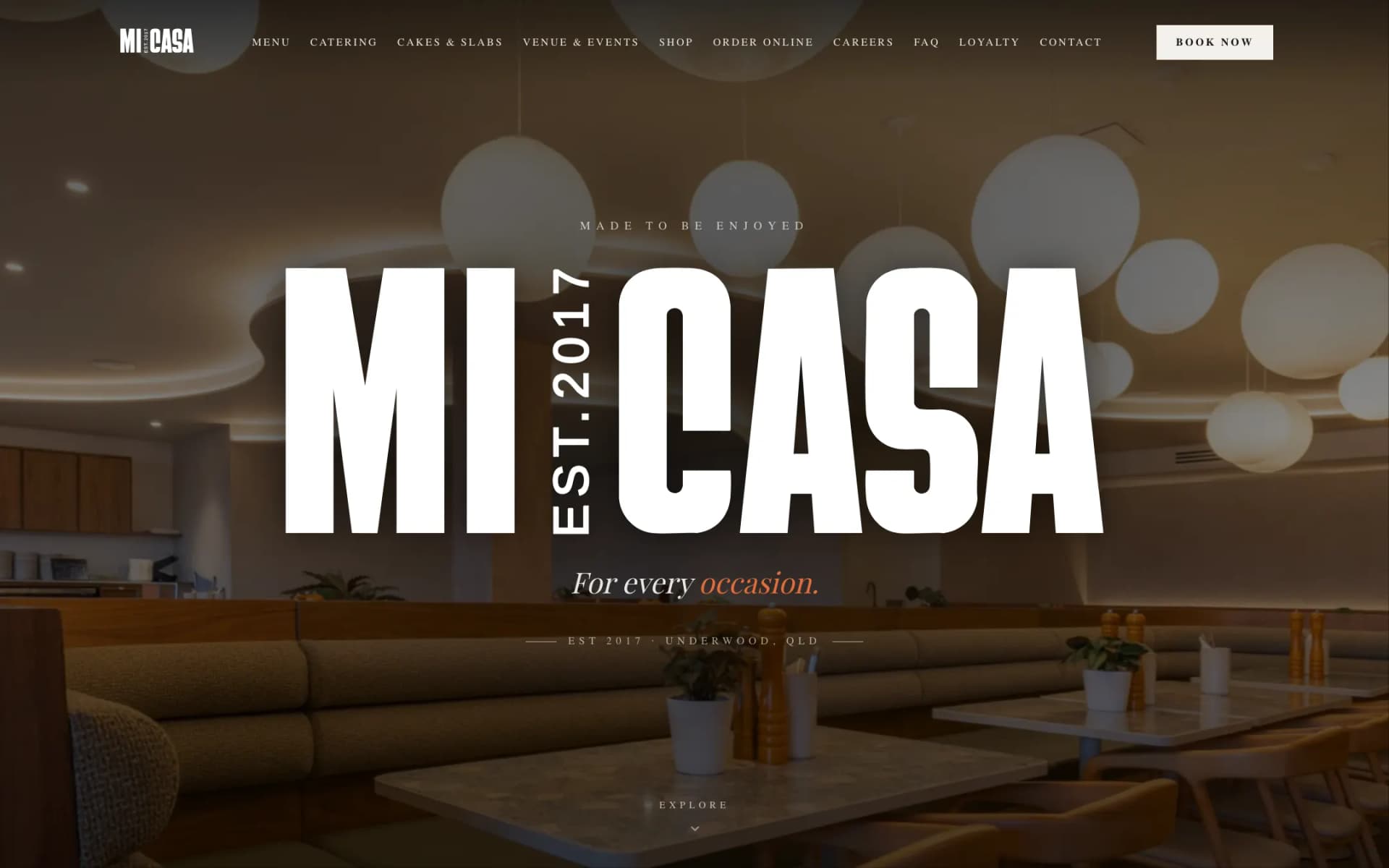 Mi Casa Cafe homepage hero: a giant white condensed MI CASA wordmark with a vertical EST.2017 between the words, over a dimmed photo of the Underwood dining room with globe pendant lights and booth seating, the tagline 'For every occasion.' and a slim navigation bar with a Book Now button